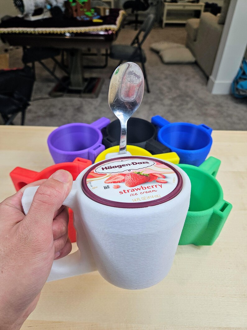 Pu&ograve; includere: Un supporto bianco a forma di tazza con manico, progettato per contenere una pinta di gelato. Il coperchio di un contenitore di gelato alla fragola &egrave; visibile, con un cucchiaio sopra. Sono visibili anche altri supporti colorati in rosso, verde, blu, giallo e viola.