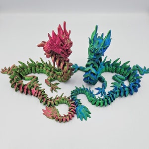 May include: Two articulated dragon figurines. One dragon is primarily green with pink accents, the other is blue and green. The dragons are posed on a white background. The dragons have detailed scales and a serpentine design.