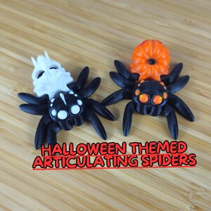 Puede incluir: Dos ara&ntilde;as articuladas con tem&aacute;tica de Halloween. Una ara&ntilde;a es negra con una cabeza de calavera blanca y la otra es negra con una cabeza de calabaza naranja.