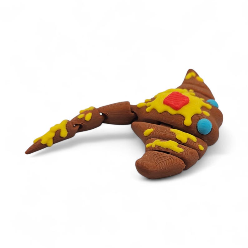 May include: A brown, toy axe with a curved blade and a long handle. The axe is decorated with yellow and red accents, and blue dots. The toy is made of a textured material and is set against a white background.
