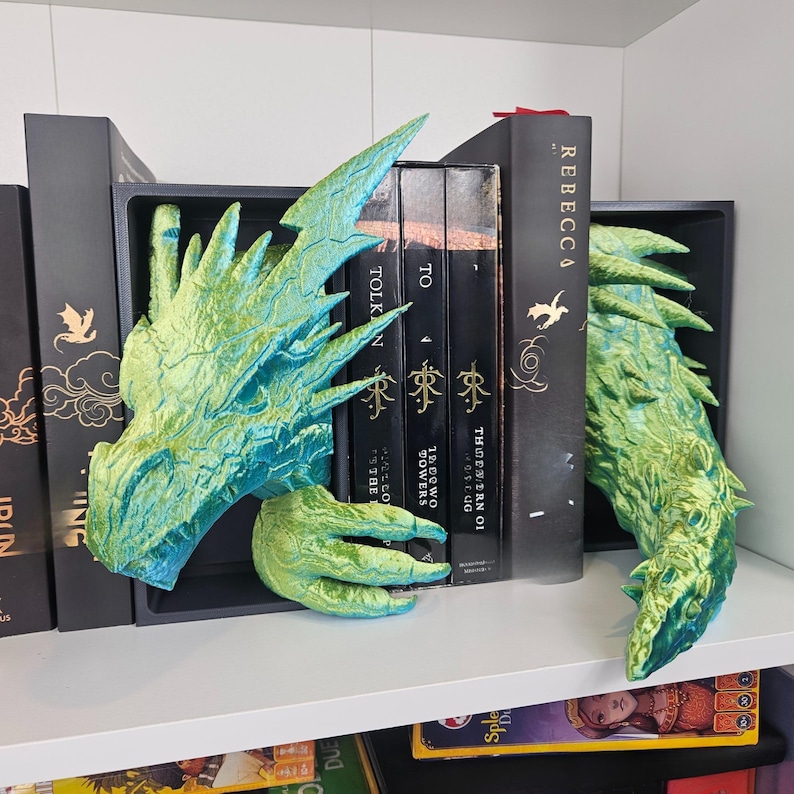 May include: Green and blue dragon-themed bookends. The bookends are made of a textured material and are designed to look like a dragon's head and tail. The bookends are on a white shelf with books in between them.