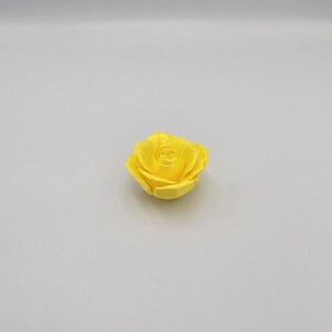 May include: A single, bright yellow rose blossom. The flower is fully open, displaying multiple layers of petals. The rose is centered on a plain, light gray background, highlighting its vibrant color and delicate form.