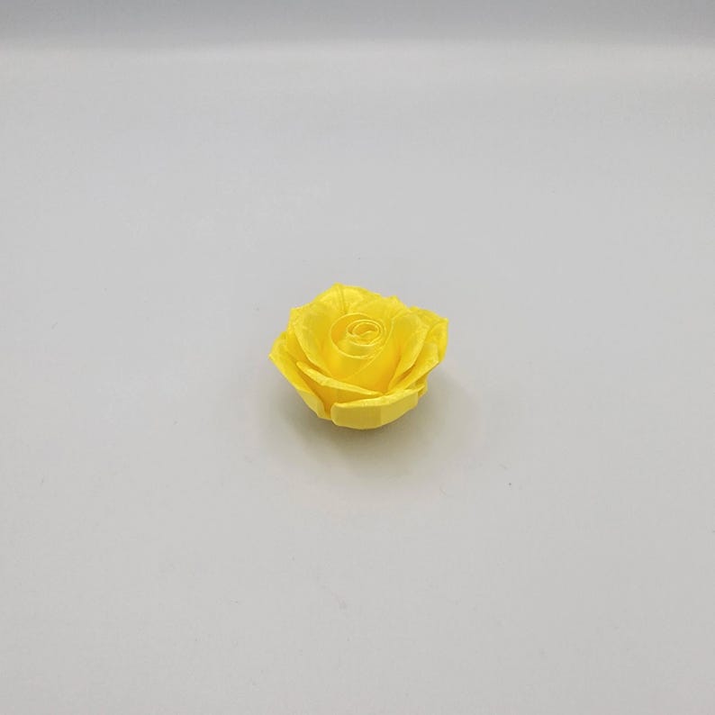 May include: A single, bright yellow rose blossom. The flower is fully open, displaying multiple layers of petals. The rose is centered on a plain, light gray background, highlighting its vibrant color and delicate form.