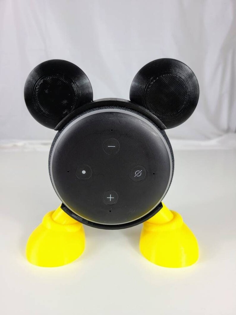 Puede incluir: Un altavoz Amazon Echo Dot negro con un dise&ntilde;o de Mickey Mouse impreso en 3D. El altavoz tiene pies amarillos y orejas negras.