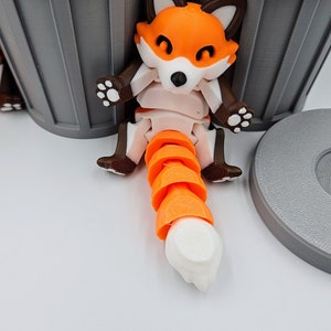 May include: A 3D-printed articulated fox figurine with orange, white, and brown coloring. The fox has a segmented orange tail and is posed near a gray trash can. The fox has a black nose and white paws.