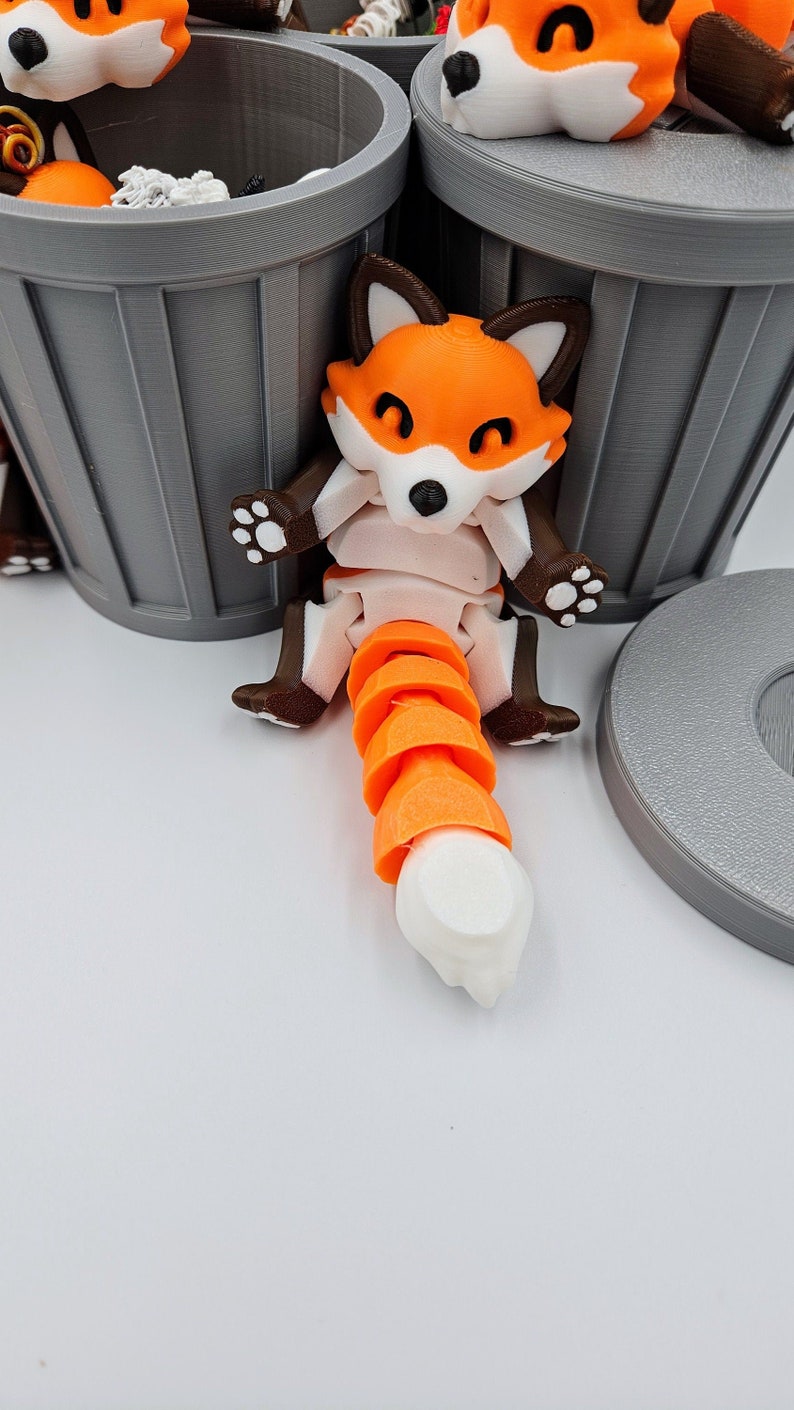 May include: A 3D-printed articulated fox figurine with orange, white, and brown coloring. The fox has a segmented orange tail and is posed near a gray trash can. The fox has a black nose and white paws.