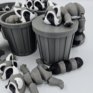 May include: A collection of gray and white articulated raccoon figures, some resting on gray trash cans. The raccoons have black and white facial markings and are in various poses, with some disassembled.