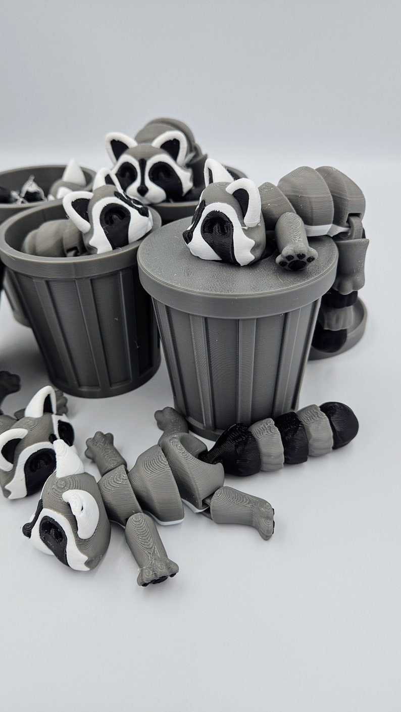 May include: A collection of gray and white articulated raccoon figures, some resting on gray trash cans. The raccoons have black and white facial markings and are in various poses, with some disassembled.