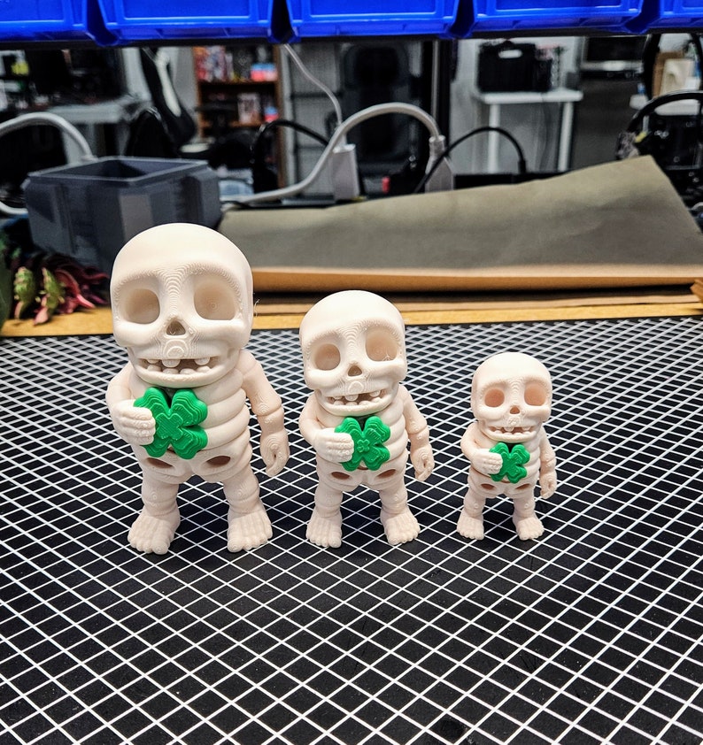 May include: Three white skeleton figurines holding green shamrock charms. The skeletons are different sizes, with the largest one on the left and the smallest one on the right.