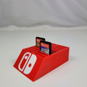 Może przedstawiać: Czerwony plastikowy stojak na karty do gier Nintendo Switch z białym logo Nintendo Switch. Stojak ma 16 miejsc na karty do gier i jest zaprojektowany tak, aby karty stały pionowo. W stojaku widoczne są dwie karty do gier, jedna do gry "The Legend of Zelda: Breath of the Wild" i jedna do gry "Super Mario Odyssey".