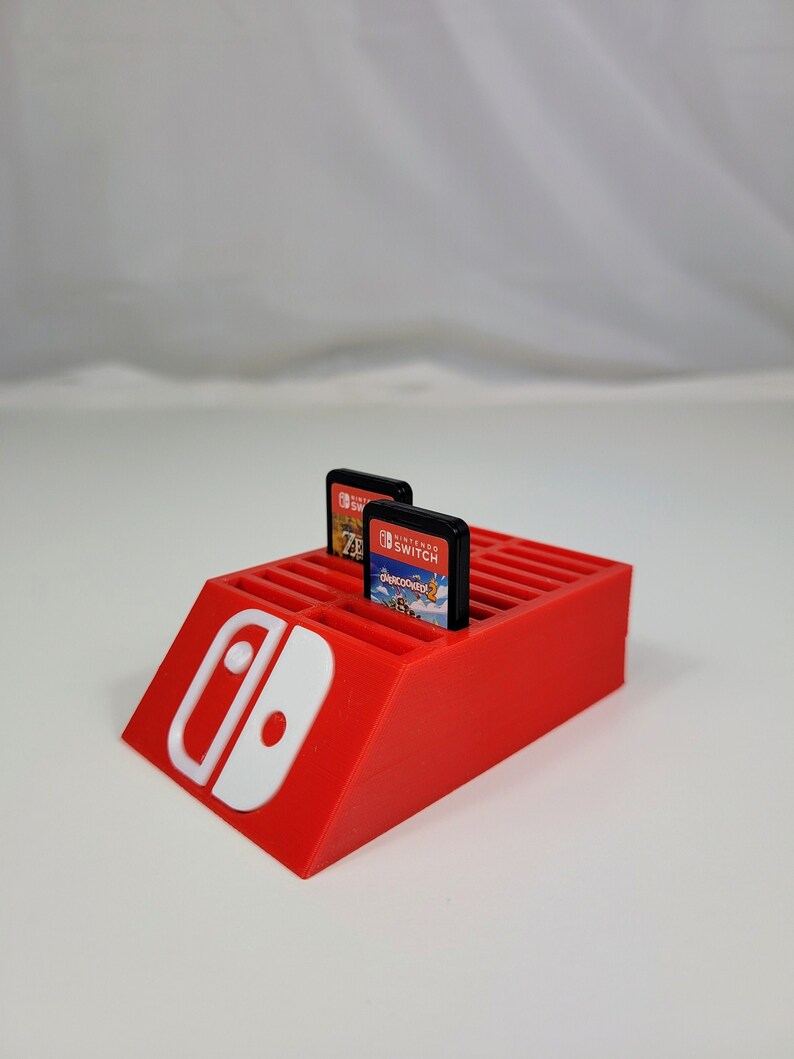Może przedstawiać: Czerwony plastikowy stojak na karty do gier Nintendo Switch z białym logo Nintendo Switch. Stojak ma 16 miejsc na karty do gier i jest zaprojektowany tak, aby karty stały pionowo. W stojaku widoczne są dwie karty do gier, jedna do gry "The Legend of Zelda: Breath of the Wild" i jedna do gry "Super Mario Odyssey".