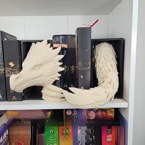 May include: A beige dragon head and tail sculpture. The head features detailed scales and a sharp crest. The tail has a feathered design. The sculpture is displayed on a white shelf with books in the background.