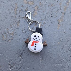 Pode incluir: Um chaveiro de boneco de neve branco com um chap&eacute;u preto, uma echarpe vermelha e bra&ccedil;os de galho castanho. O chaveiro tem um anel de metal prateado e um clipe prateado.