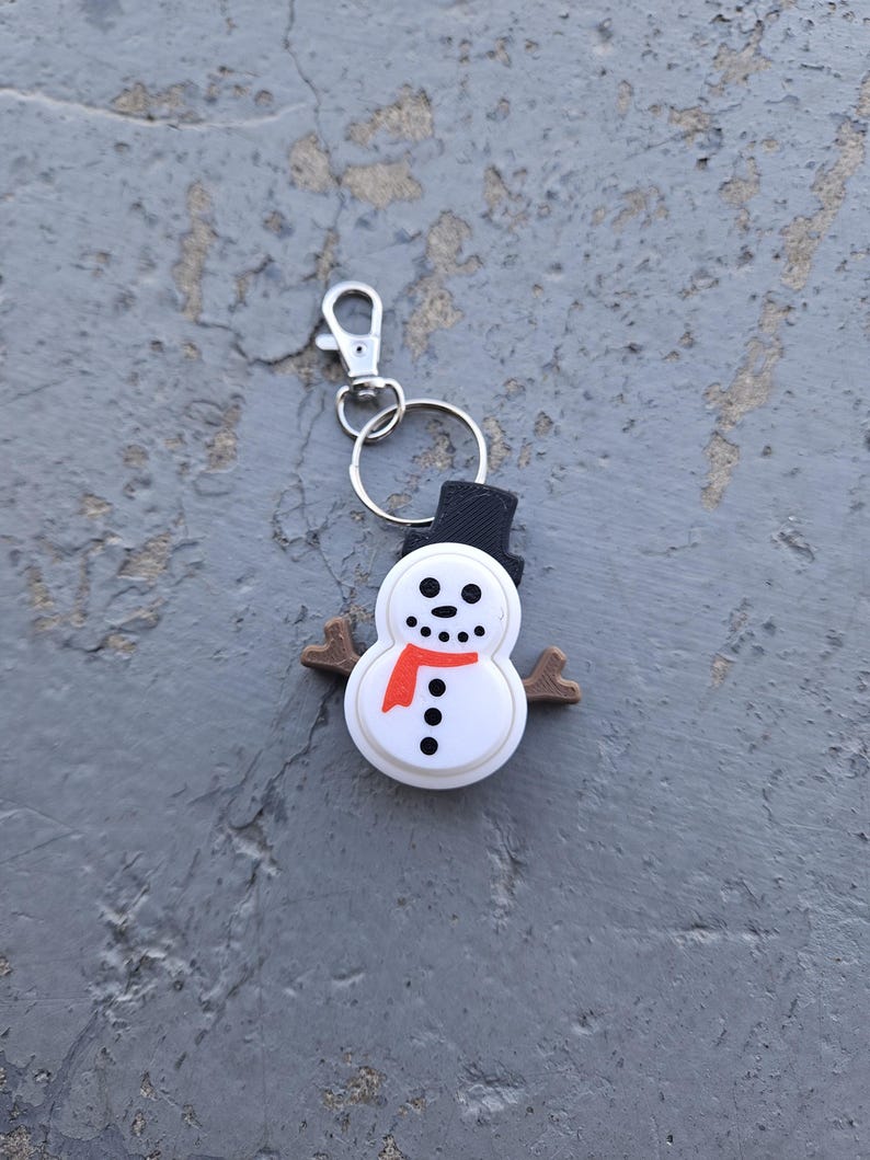 Pode incluir: Um chaveiro de boneco de neve branco com um chap&eacute;u preto, uma echarpe vermelha e bra&ccedil;os de galho castanho. O chaveiro tem um anel de metal prateado e um clipe prateado.
