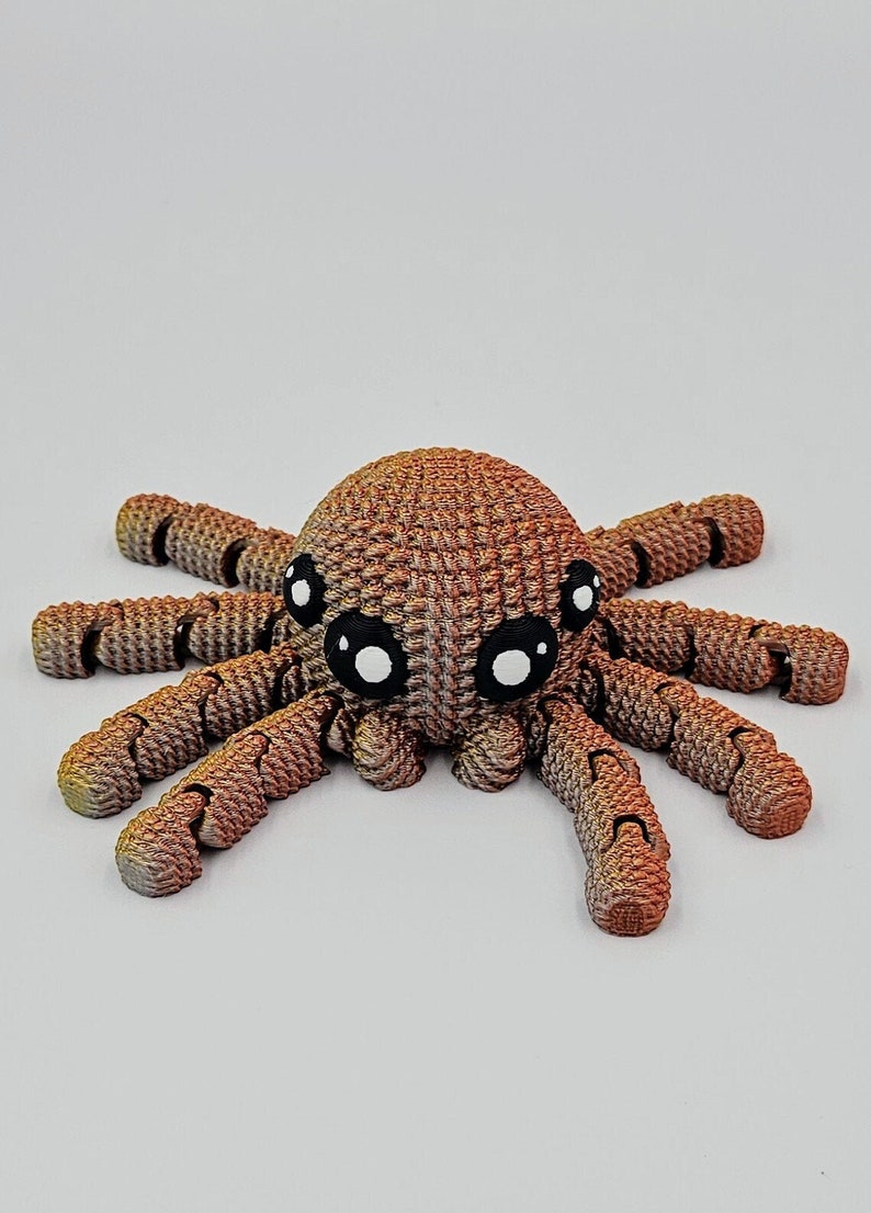 Op de afbeelding: Een 3D-geprint speelgoedspin in bruin en oranje met grote zwarte ogen. De spin heeft acht poten en een textuurrijk lichaam.