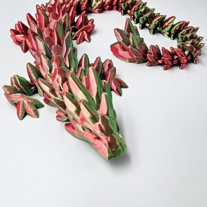 K&ouml;nnte beinhalten: Eine 3D-gedruckte Drachenkette aus rotem und gr&uuml;nem Kunststoff. Der Drache ist zu einem Kreis zusammengerollt, mit dem Kopf auf dem Schwanz. Die Kette besteht aus einzelnen Schuppen, die miteinander verbunden sind.