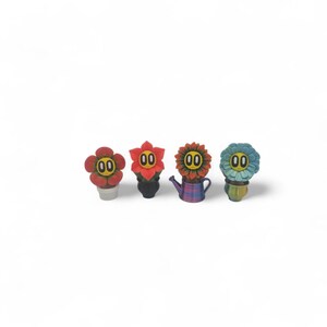 May include: Four whimsical flower-shaped figurines with smiling faces and the number "00" on their faces. The flowers are red, orange, and blue, and are in small pots or a watering can. The background is white.