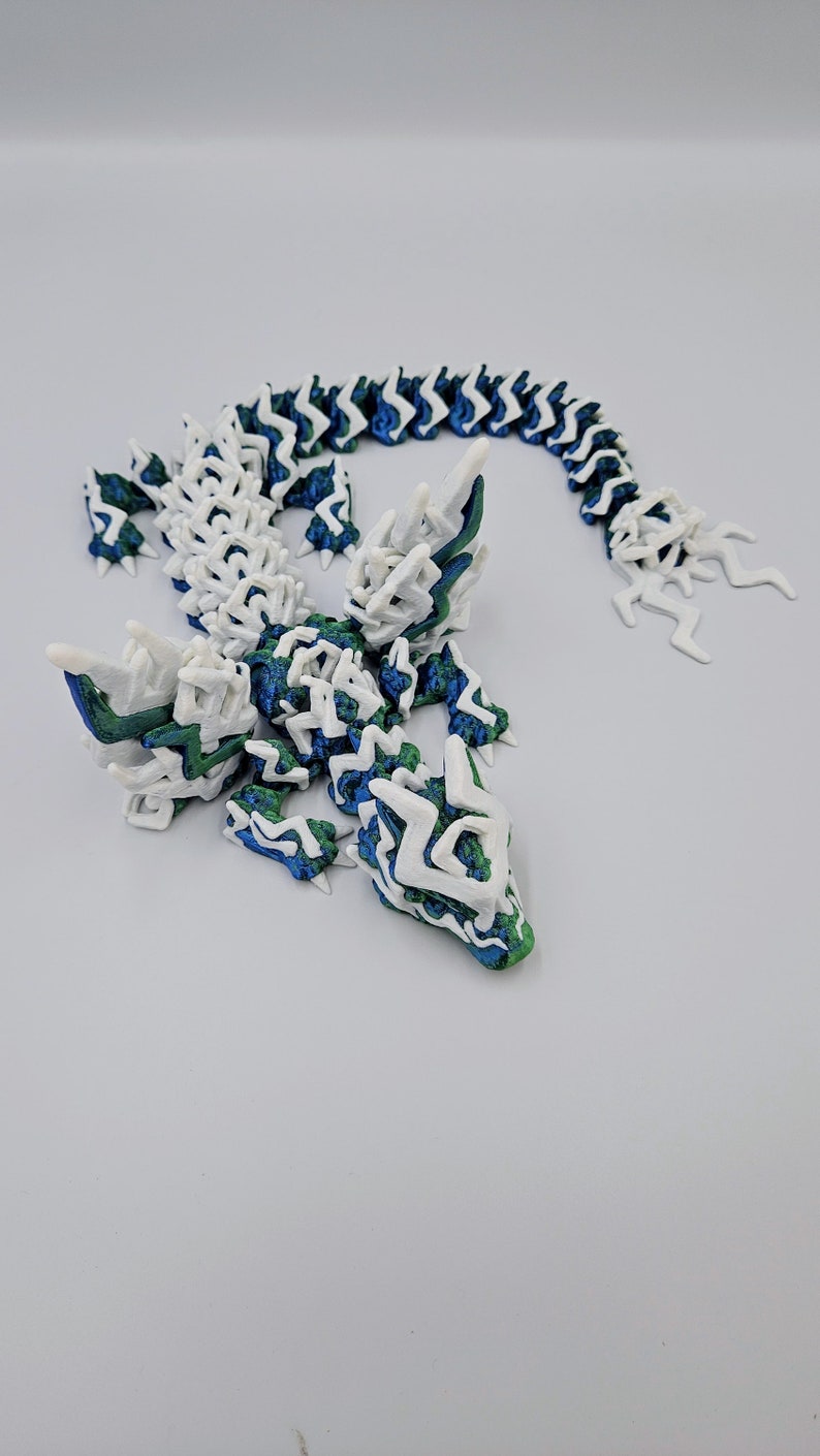 May include: A 3D printed lizard sculpture with a white, blue and green colour scheme. The lizard is posed in a standing position with its tail curled up behind it.