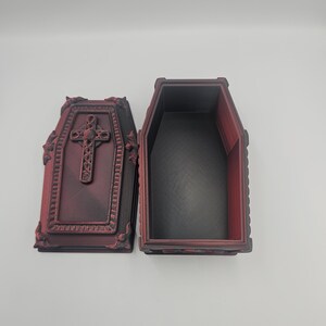 May include: An open, decorative coffin with a cross on the lid. The coffin is dark red with black interior and ornate detailing. The lid is open, revealing the inside of the coffin.