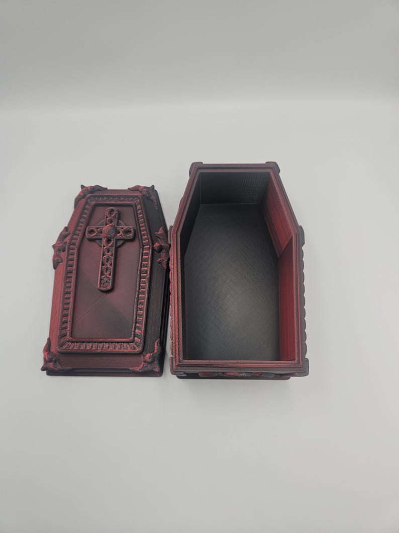 May include: An open, decorative coffin with a cross on the lid. The coffin is dark red with black interior and ornate detailing. The lid is open, revealing the inside of the coffin.