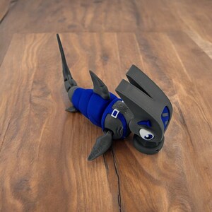 May include: A blue and gray 3D printed toy shaped like a hammerhead shark. The shark has a large, flat head with a wide mouth and large, round eyes. The shark is wearing a blue and gray suit with a belt.