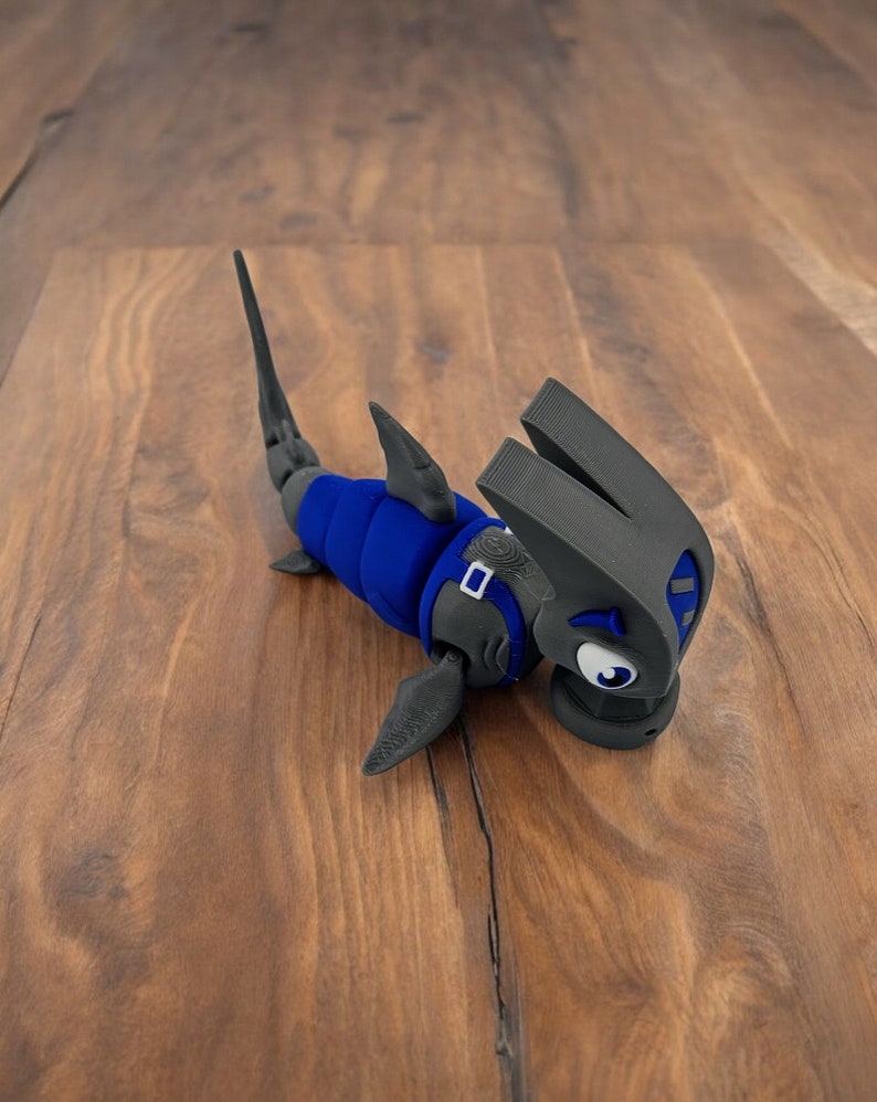 May include: A blue and gray 3D printed toy shaped like a hammerhead shark. The shark has a large, flat head with a wide mouth and large, round eyes. The shark is wearing a blue and gray suit with a belt.