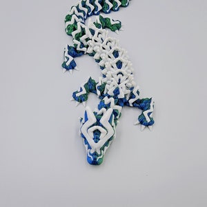 May include: A white and blue 3D printed lizard with a green and blue patterned design. The lizard is posed in a standing position with its head tilted up.