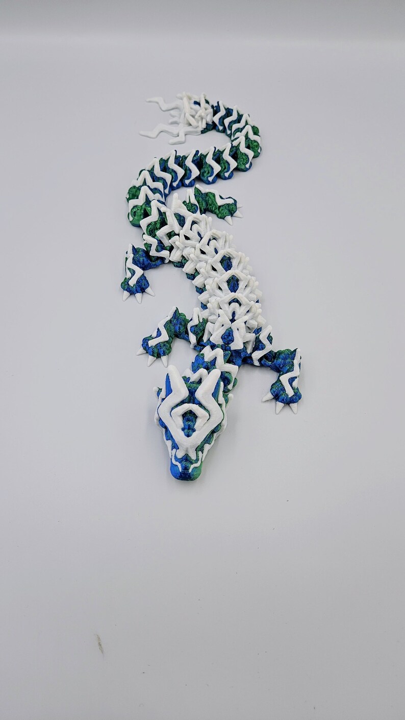 May include: A white and blue 3D printed lizard with a green and blue patterned design. The lizard is posed in a standing position with its head tilted up.