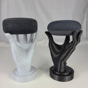 Op de afbeelding: Twee 3D-geprinte handen die elk een zwarte slimme speaker vasthouden. De handen staan op een witte en zwarte basis.