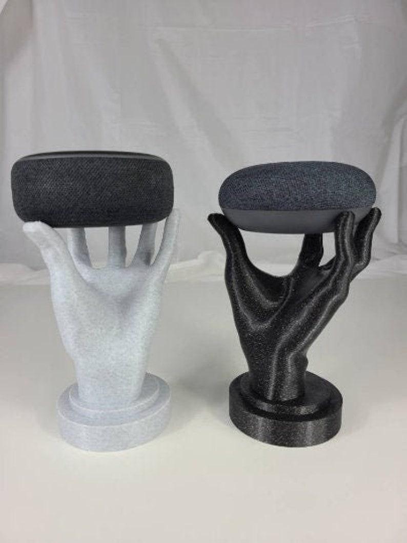 Op de afbeelding: Twee 3D-geprinte handen die elk een zwarte slimme speaker vasthouden. De handen staan op een witte en zwarte basis.