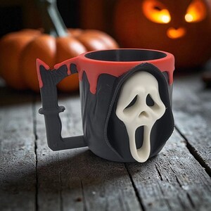 Pode incluir: Caneca preta e vermelha com tema de Halloween com um design de m&aacute;scara Ghostface branca. A al&ccedil;a tem formato de faca. A caneca tem um efeito de tinta vermelha escorrendo ao redor da borda. Ab&oacute;boras est&atilde;o no fundo.