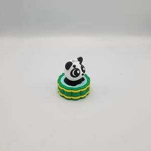May include: A small, panda-shaped figurine with a black and white body, perched atop a green and yellow tiered base. The panda has large, expressive eyes and black ears. The base has a scalloped edge.