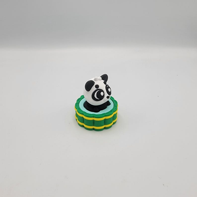 May include: A small, panda-shaped figurine with a black and white body, perched atop a green and yellow tiered base. The panda has large, expressive eyes and black ears. The base has a scalloped edge.