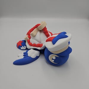 Pode incluir: Uma estatueta azul, branca e vermelha com chap&eacute;u de marinheiro e design de lua crescente. A estatueta tem um chap&eacute;u branco com aba azul e uma parte superior listrada em vermelho e branco. A estatueta est&aacute; deitada de lado.