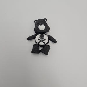 May include: A black bear figurine with a white belly featuring a skull and crossbones design. The bear has small black ears, arms, and legs. The figurine is set against a plain white background.