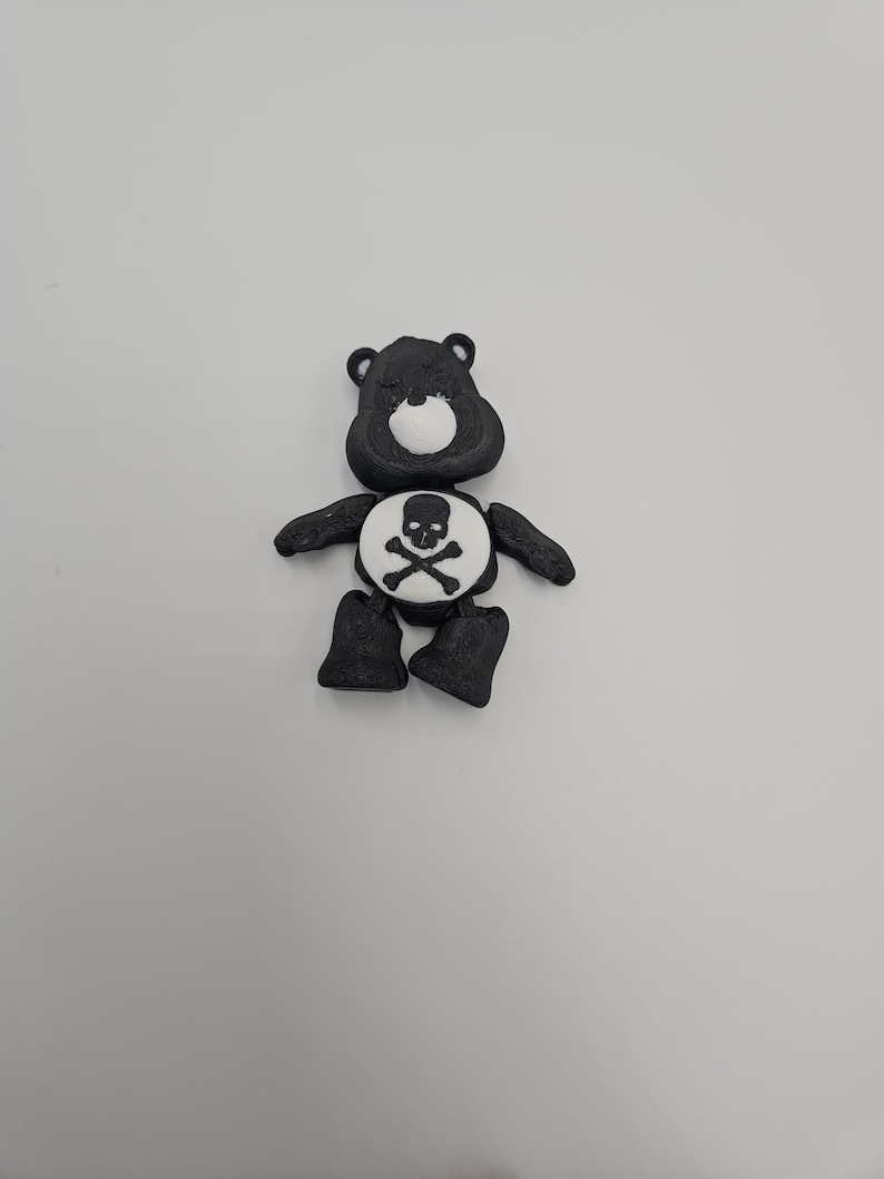 May include: A black bear figurine with a white belly featuring a skull and crossbones design. The bear has small black ears, arms, and legs. The figurine is set against a plain white background.