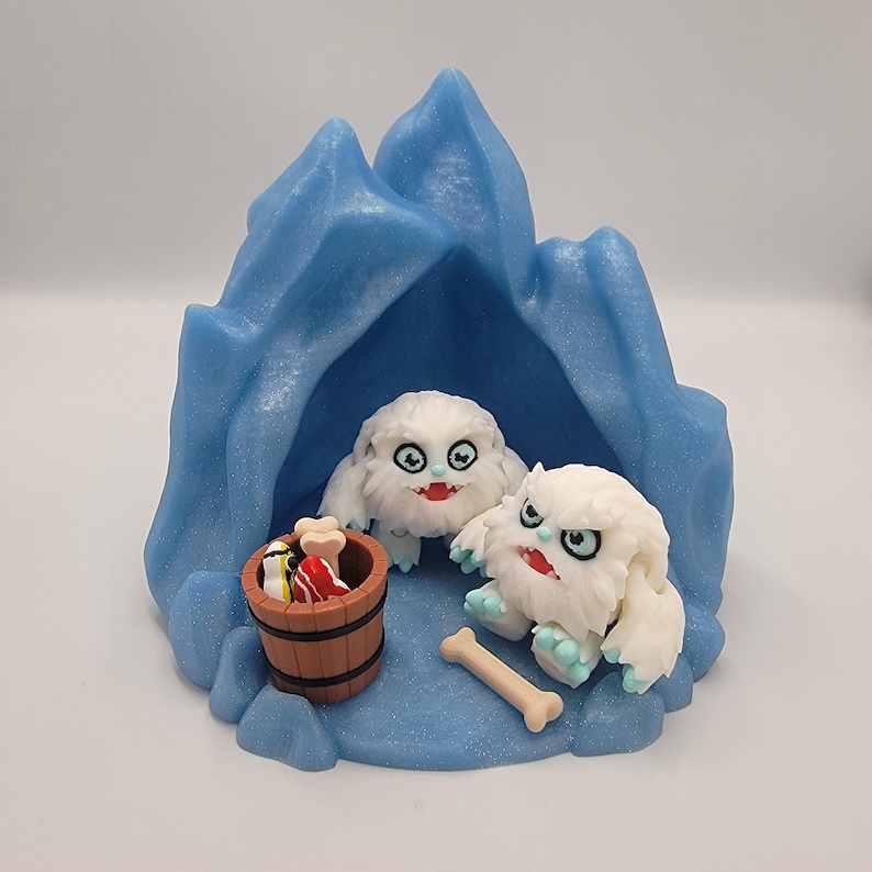 May include: A whimsical diorama featuring two white, furry yeti figures with blue accents, set inside a blue ice cave. A small wooden barrel holds colorful items, and a bone lies nearby. The scene is set on a light blue base.