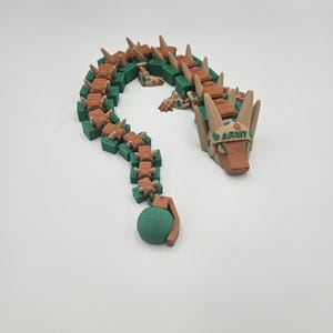 Pode incluir: Um brinquedo de drag&atilde;o articulado impresso em 3D em tons de castanho, verde e bege. O drag&atilde;o tem um corpo segmentado, uma cabe&ccedil;a com a palavra "ARMY" impressa e uma granada verde na ponta da cauda. O brinquedo &eacute; projetado para ser flex&iacute;vel e pos&aacute;vel.