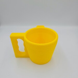 Pu&ograve; includere: Una tazza giallo brillante con un'ampia maniglia rettangolare. La tazza ha un design semplice e moderno e una finitura liscia e opaca. Sembra essere fatta di plastica o un materiale simile. Lo sfondo &egrave; bianco.