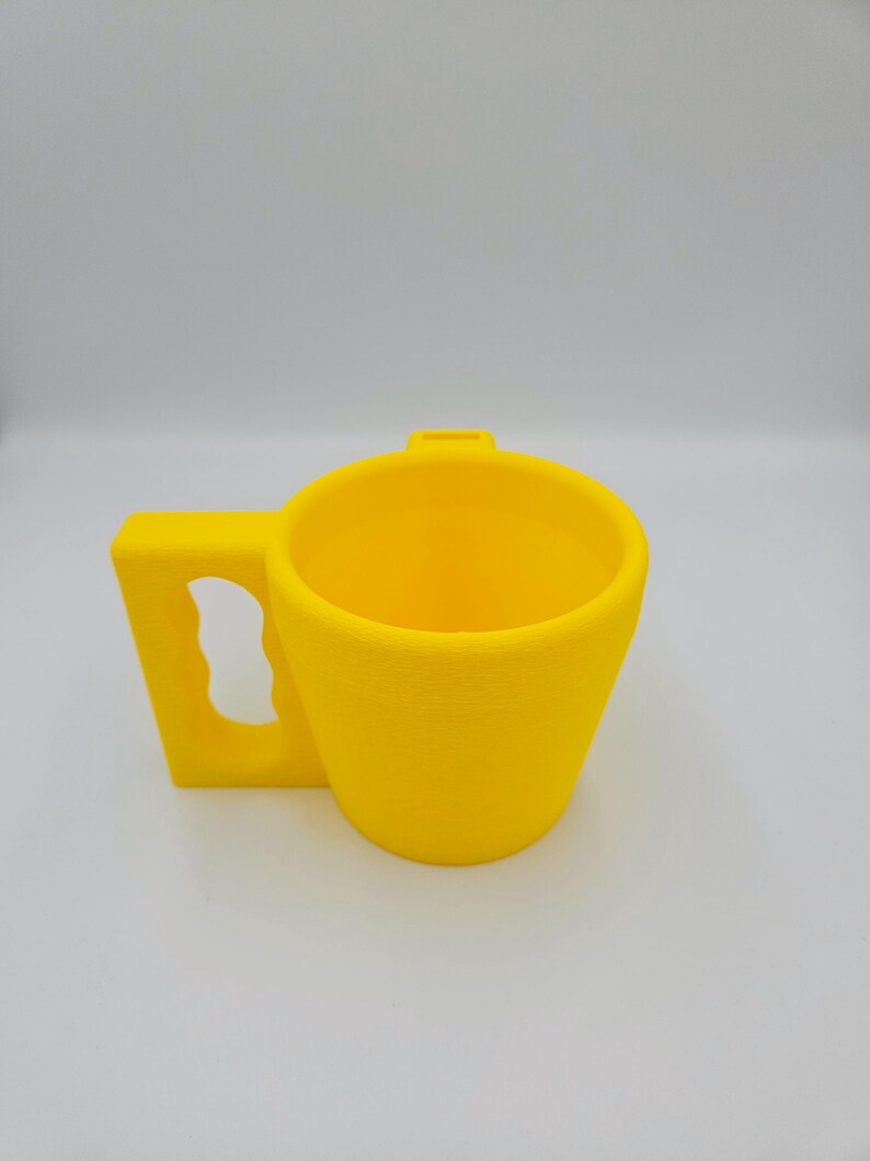 Pu&ograve; includere: Una tazza giallo brillante con un'ampia maniglia rettangolare. La tazza ha un design semplice e moderno e una finitura liscia e opaca. Sembra essere fatta di plastica o un materiale simile. Lo sfondo &egrave; bianco.