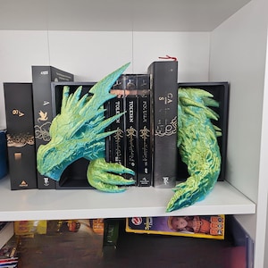 May include: A pair of dragon-themed bookends in shades of green and blue. The bookends are designed to look like a dragon's head and tail, and are placed on a white shelf with books.
