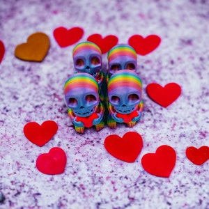 May include: Five small, colourful, rainbow-painted skull figurines holding red heart-shaped objects. The figurines are arranged in a row on a light purple background with red hearts scattered around them.
