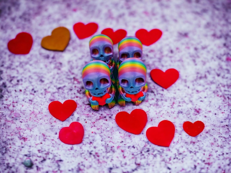 May include: Five small, colourful, rainbow-painted skull figurines holding red heart-shaped objects. The figurines are arranged in a row on a light purple background with red hearts scattered around them.