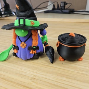 Puede incluir: Una figura de bruja de Halloween impresa en 3D con un sombrero negro, pelo naranja y un vestido morado, sosteniendo una cuchara verde y una escoba negra. Junto a ella hay un caldero negro con tapa naranja. La bruja tiene un collar.