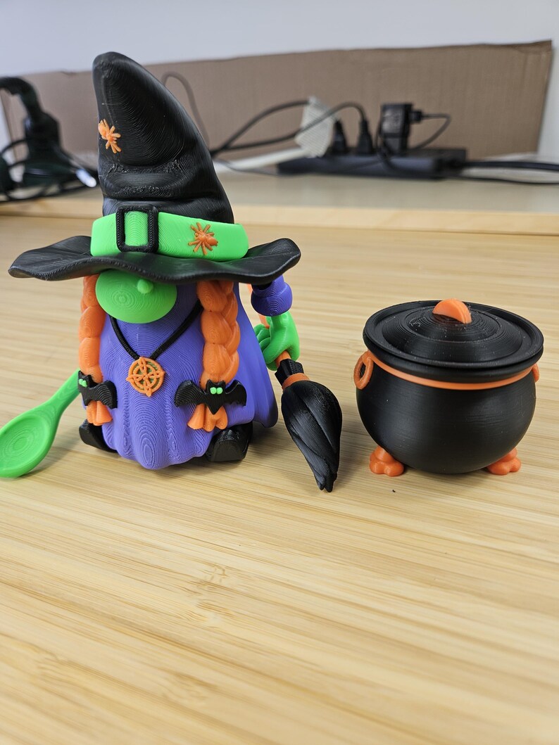 Puede incluir: Una figura de bruja de Halloween impresa en 3D con un sombrero negro, pelo naranja y un vestido morado, sosteniendo una cuchara verde y una escoba negra. Junto a ella hay un caldero negro con tapa naranja. La bruja tiene un collar.