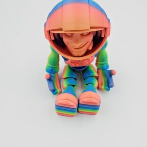 May include: A small, colorful, 3D printed figurine of a character wearing a rainbow helmet and a rainbow suit. The character is sitting with its legs crossed and has a friendly expression.