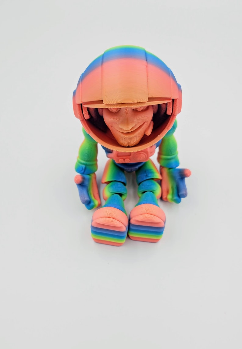 May include: A small, colorful, 3D printed figurine of a character wearing a rainbow helmet and a rainbow suit. The character is sitting with its legs crossed and has a friendly expression.