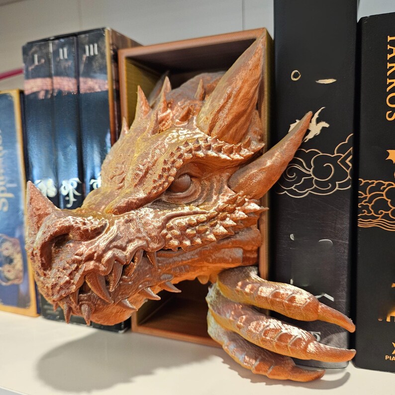 May include: A detailed, bronze-colored dragon head bookend with sharp teeth, horns, and textured scales. The dragon's claws extend from a wooden box, creating a unique and decorative bookshelf accent. The bookend is placed between several books.
