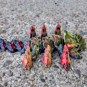 May include: A 3D printed model of a creature with a segmented body, green and black scales, and red spikes. The creature has a large head with a toothy mouth and a pair of eyes.