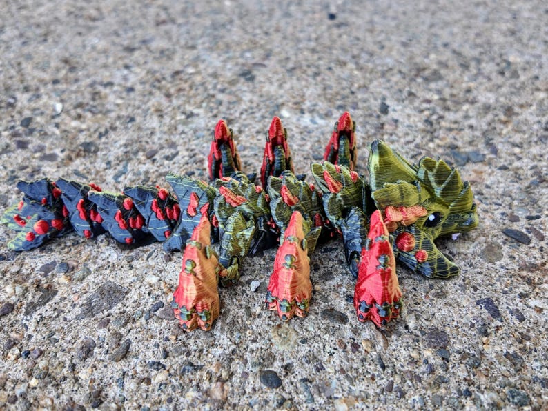May include: A 3D printed model of a creature with a segmented body, green and black scales, and red spikes. The creature has a large head with a toothy mouth and a pair of eyes.